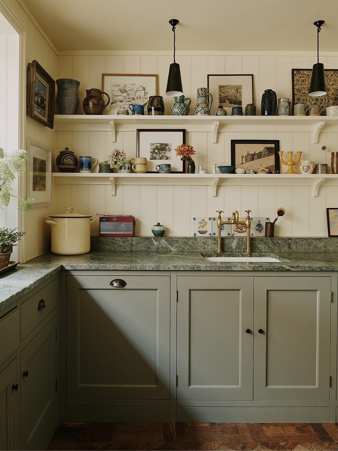 A kitchen with dark green cabinets and light wooden open shelving featuring a small landscape painting tucked between objects.