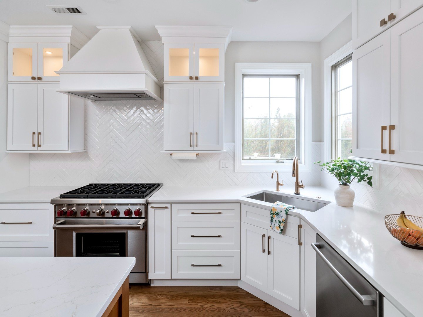 White contemporary kitchen with white shaker cabinets, brushed bronze hardware, a white chevron tile backsplash, a stainless steel range, and a window over a corner sink.