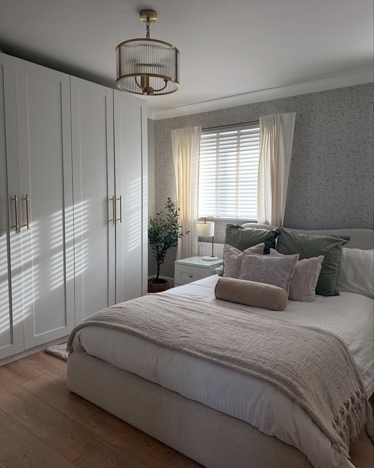A small beige and sage green bedroom featuring floor-to-ceiling white wardrobe units, patterned wallpaper behind the bed, a textured throw, and a gold geometric light fixture.