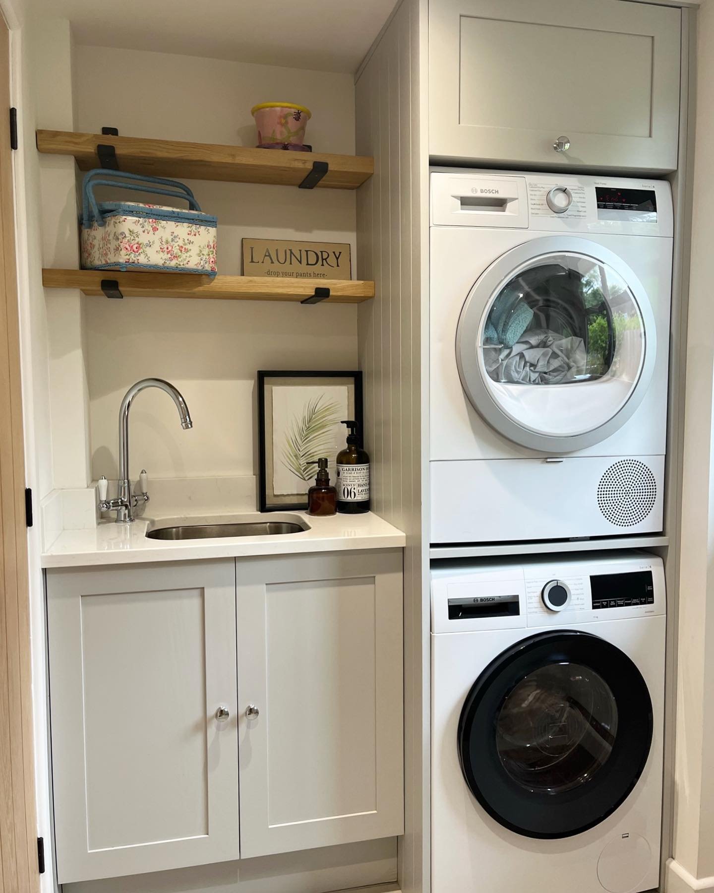 A tiny laundry nook with a stacked washer and dryer next to a small cabinet and stainless steel utility sink, finished with wooden floating shelves and a light gray color palette.