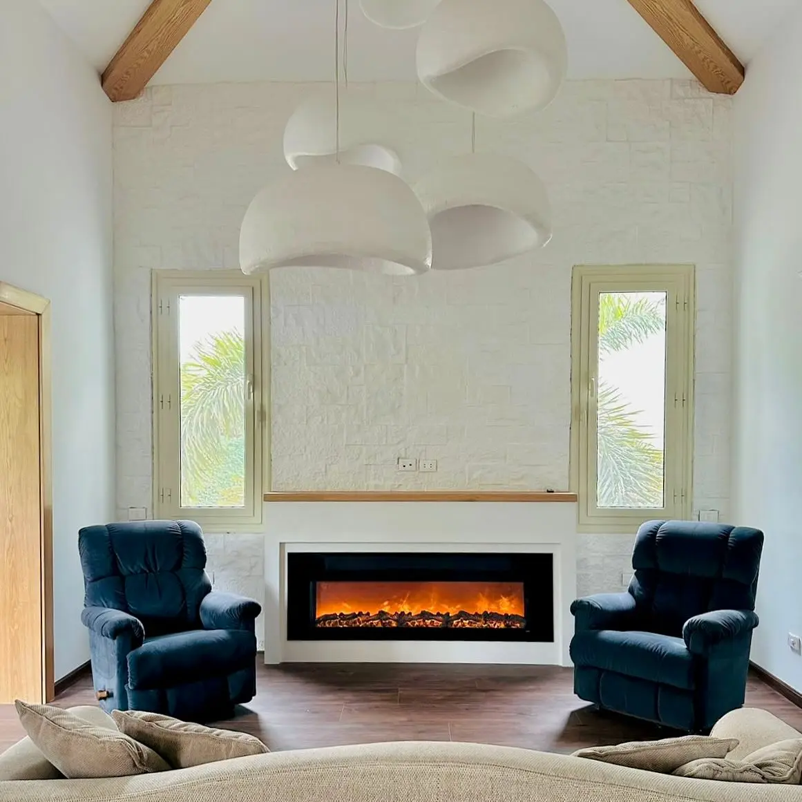 Wide, horizontal electric fireplace integrated into a white feature wall with a small floating mantel, flanked by two dark blue armchairs and tall windows, under a vaulted ceiling.