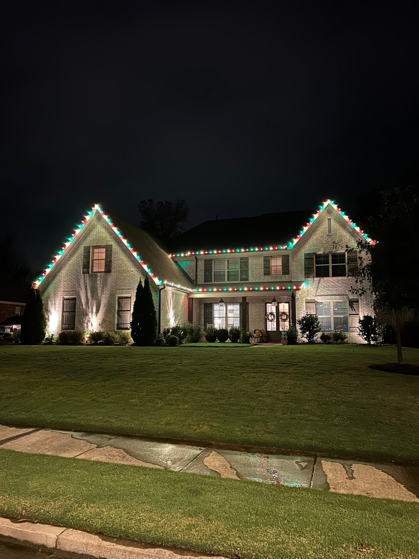 A large brick house is outlined with traditional red and green string lights along the rooflines and gutters, complemented by white floodlights highlighting the front gables.