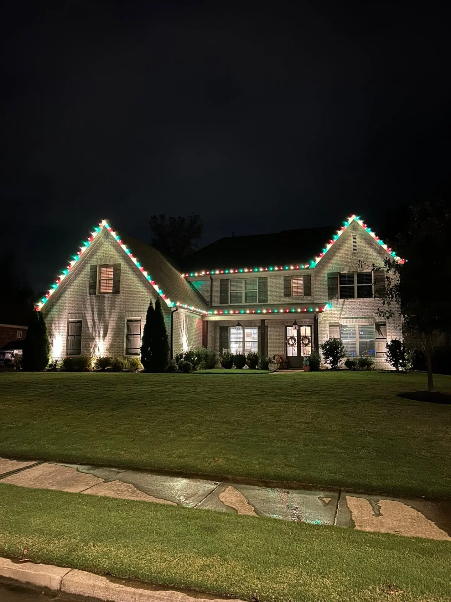 A large brick house is outlined with traditional red and green string lights along the rooflines and gutters, complemented by white floodlights highlighting the front gables.