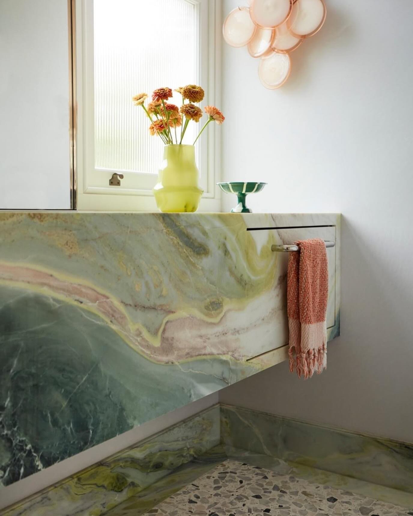 A sleek, silver recessed towel bar mounted directly beneath a dramatic green and pink marble vanity, holding a fringed hand towel.