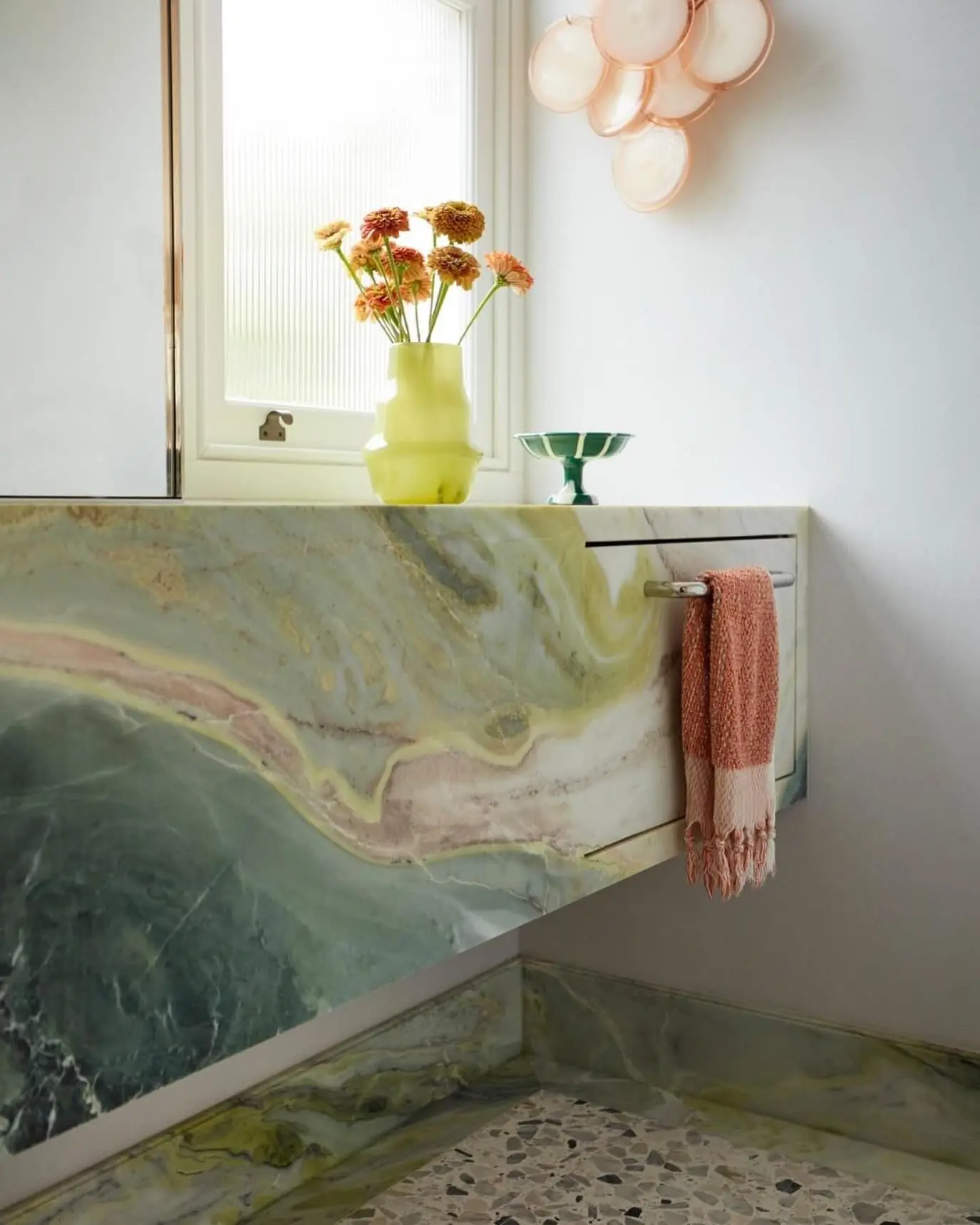 A sleek, silver recessed towel bar mounted directly beneath a dramatic green and pink marble vanity, holding a fringed hand towel.