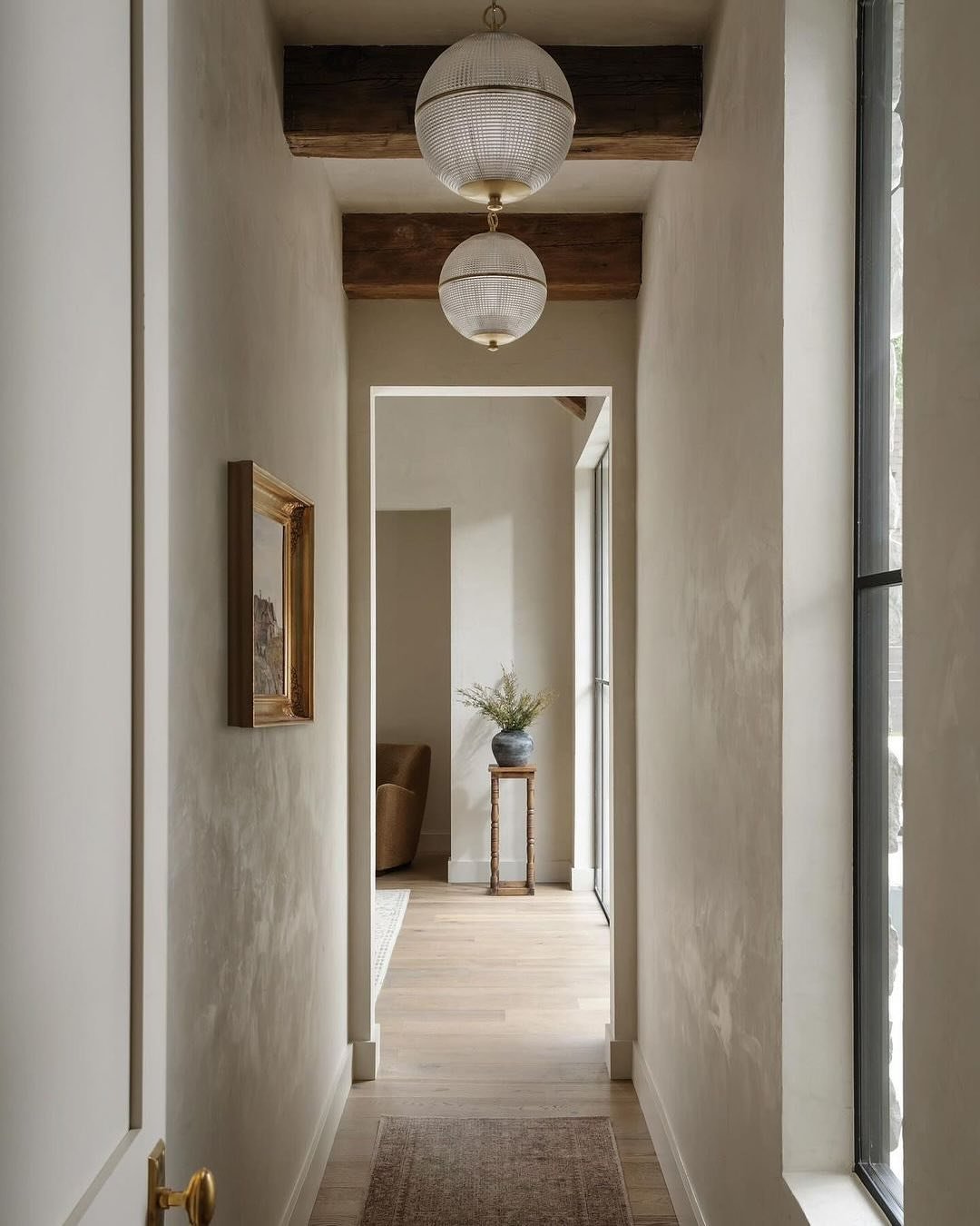 Narrow, neutral-toned hallway with light wood floors, rustic exposed wood ceiling beams, and two spherical, textured white glass pendant lights.