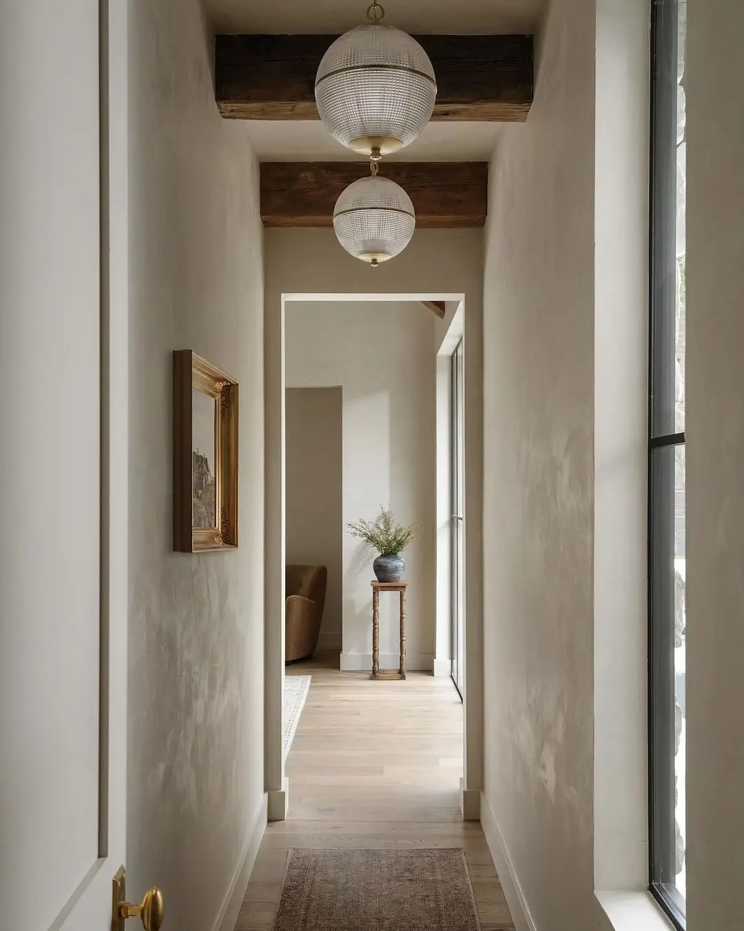 Narrow, neutral-toned hallway with light wood floors, rustic exposed wood ceiling beams, and two spherical, textured white glass pendant lights.