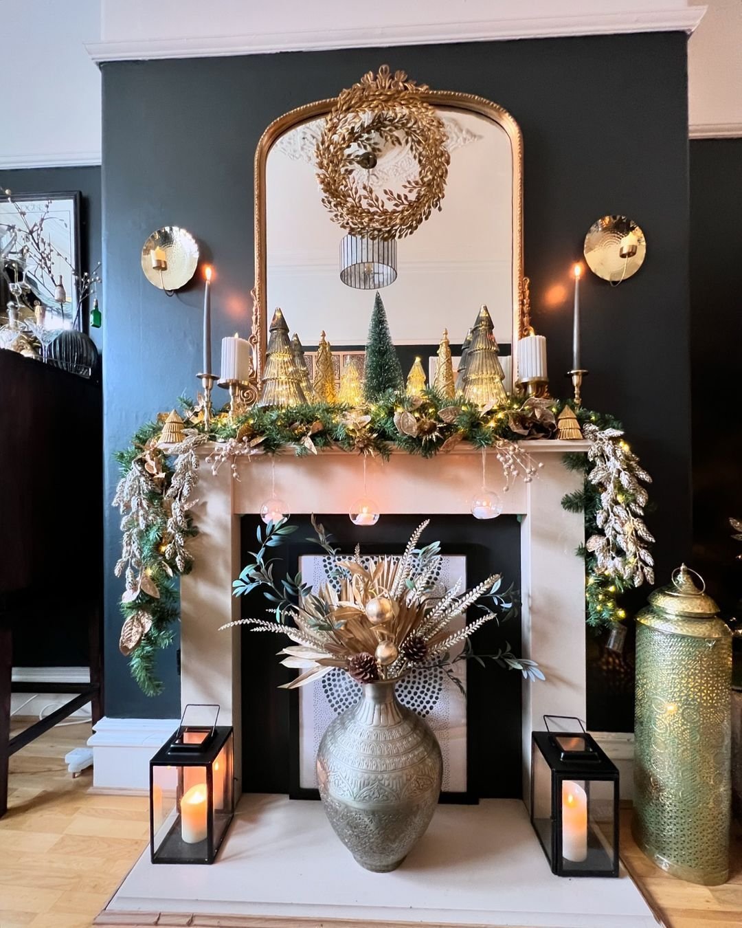 White mantel against a dark green wall, featuring an arched gold mirror, a lush garland with gold accents, and a collection of shimmering glass Christmas trees.