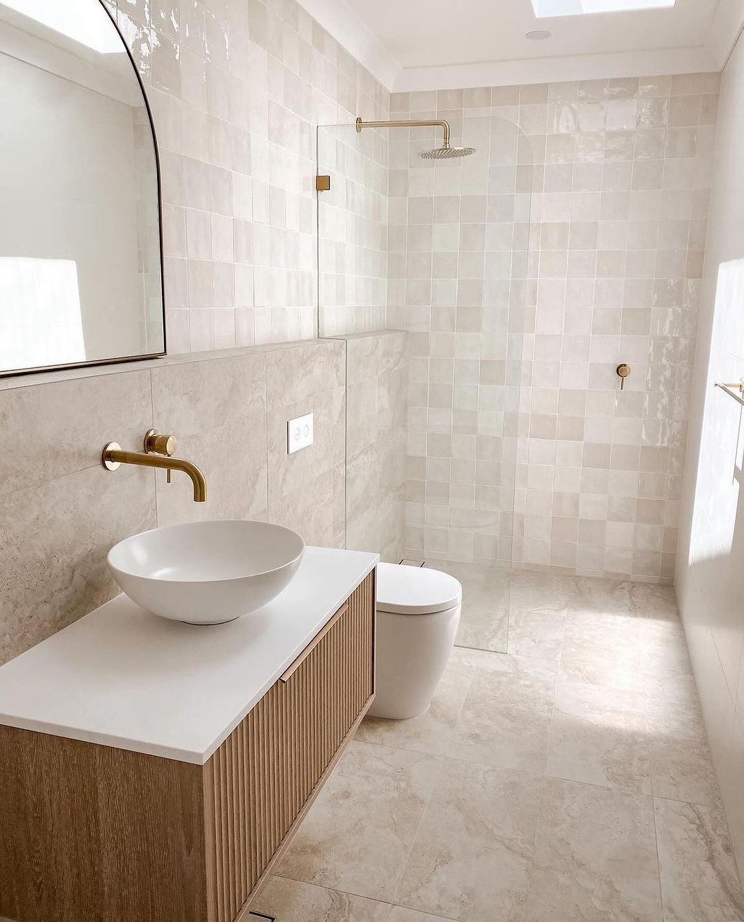 A bright, minimalist bathroom with a light wood vanity, white vessel sink, and an overall neutral palette featuring large, matte stone-look tiles on the floor.