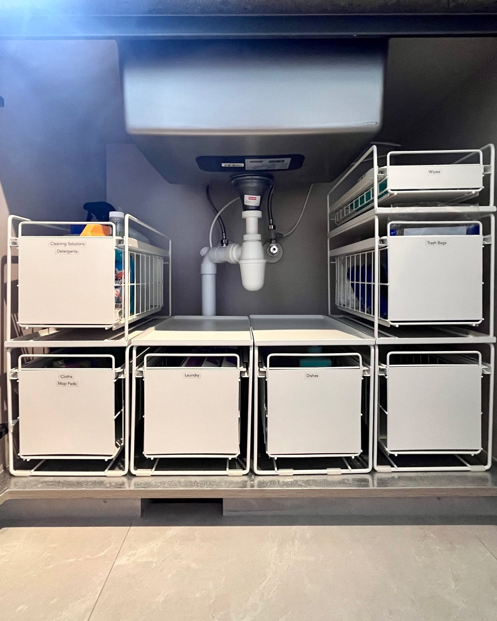 Organized cabinet under a sink with white modular sliding drawers and wire baskets around the central plumbing.