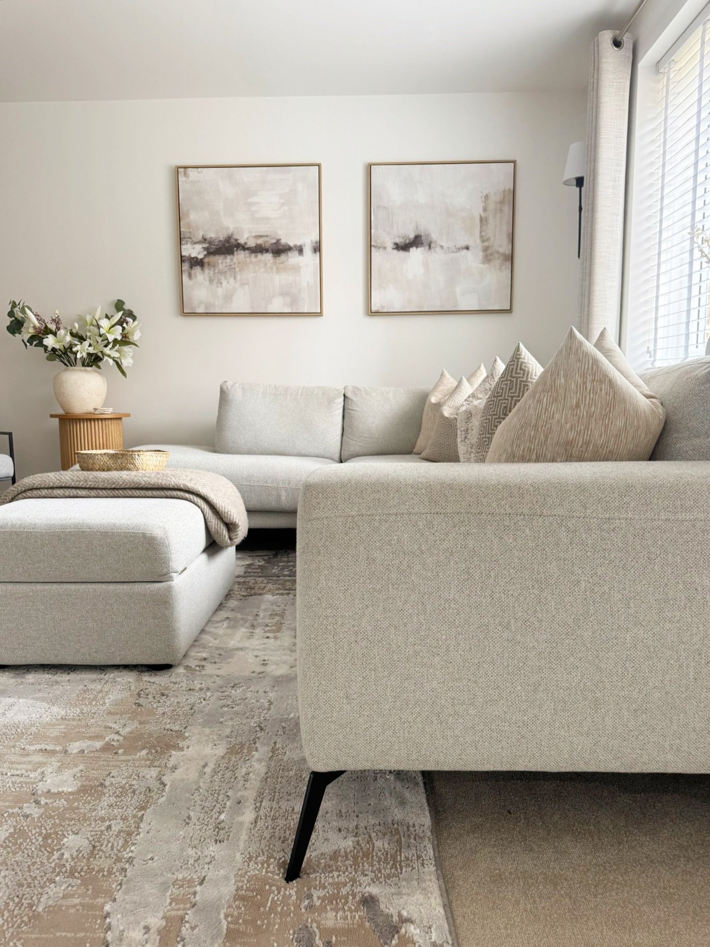 A close-up view of a light grey sectional sofa with a matching ottoman, accented by two square abstract paintings in neutral tones.