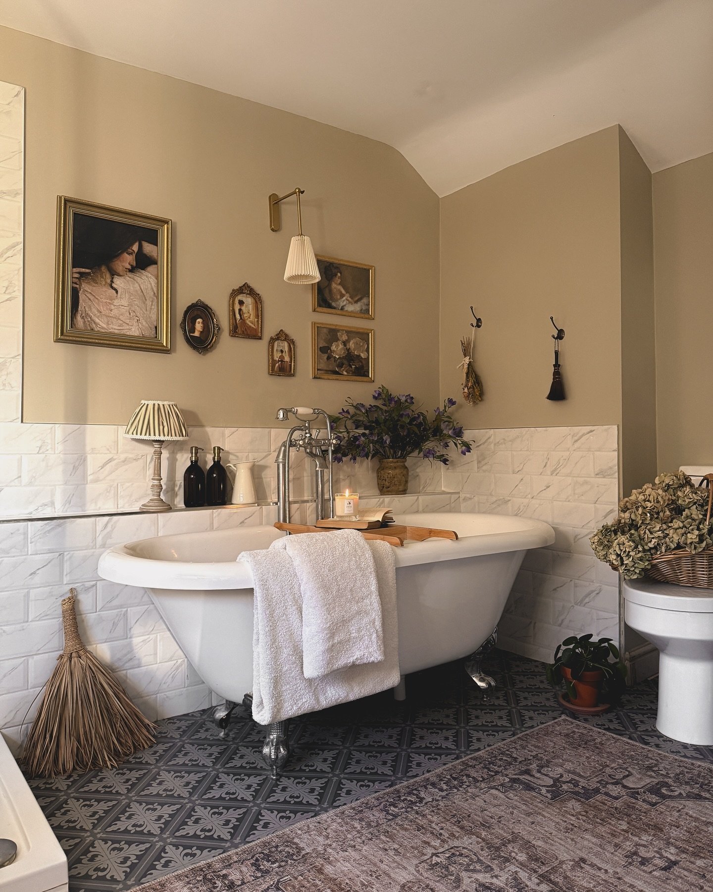 A warm beige and white bathroom featuring a clawfoot tub, a gallery wall of framed art, and a rectangular distressed gray and mauve Oriental-style rug placed over patterned blue and white floor tiles.