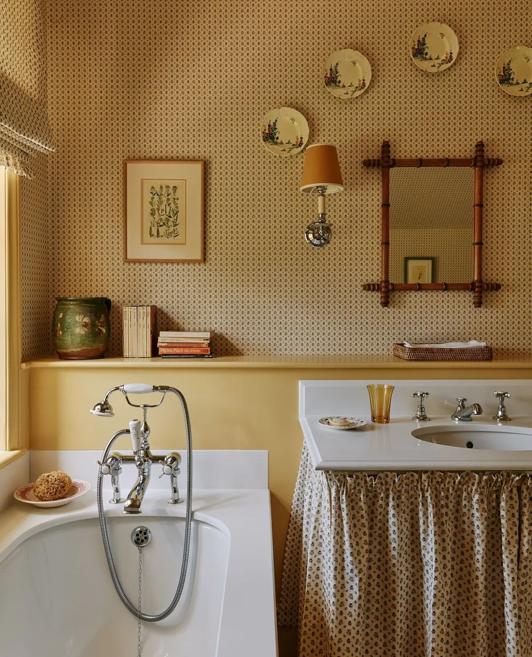 A traditional bathroom with patterned tan wallpaper and a skirted vanity, displaying a collection of decorative floral plates and a framed botanical print.