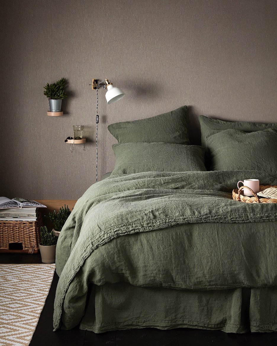 A cozy bedroom featuring a wall painted in a deep brown/taupe, with a bed completely dressed in rich, wrinkled olive green linen bedding.