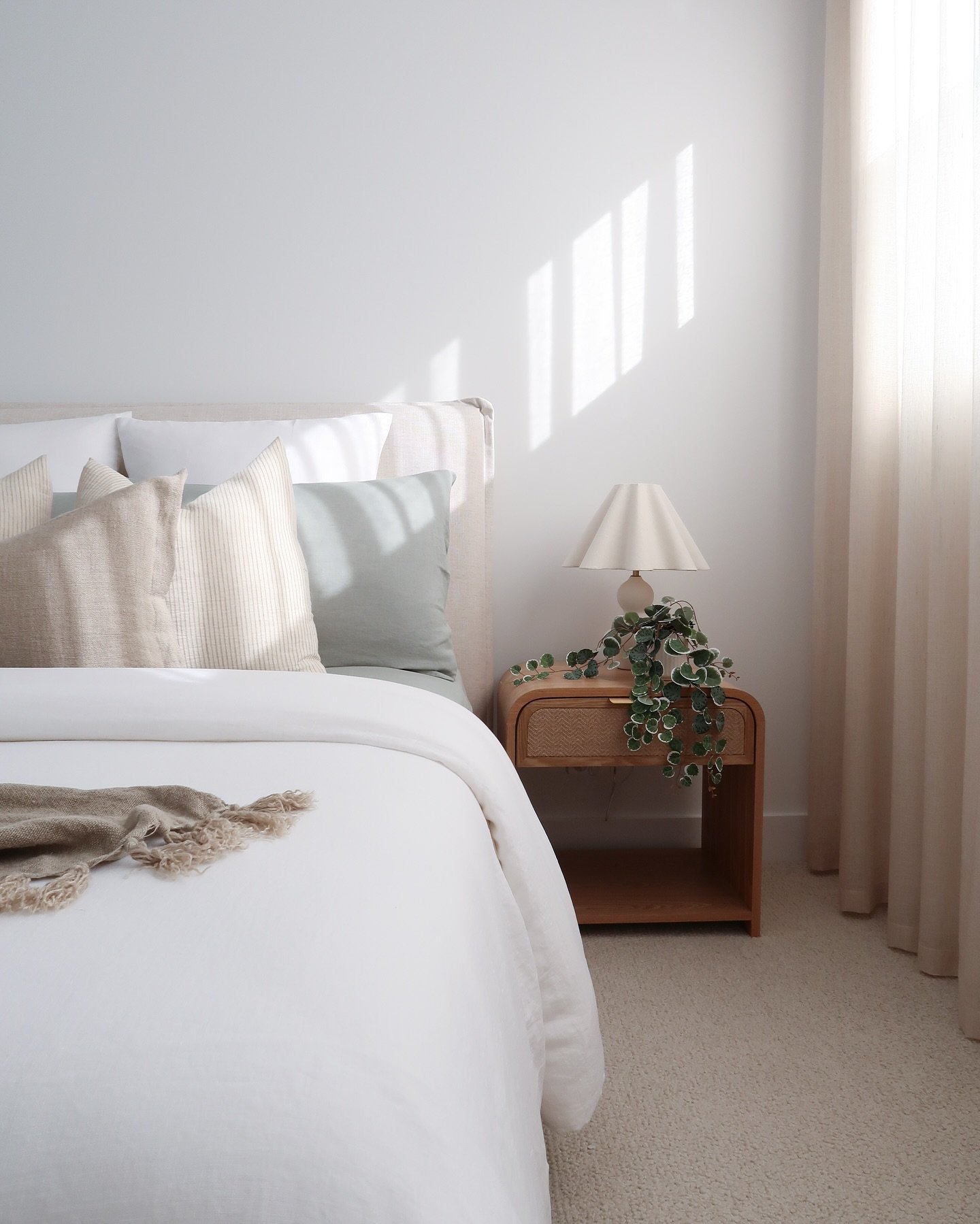 Close-up of a neutral bedroom corner featuring white bedding, striped beige and pale green cushions, a rattan nightstand, and a small trailing plant.
