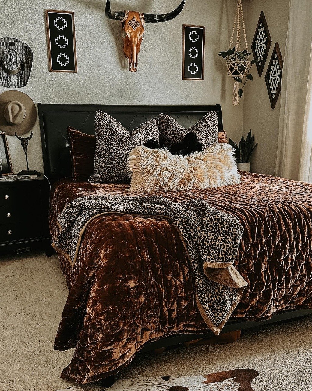 A Western bedroom featuring a burnt umber velvet quilt, leopard print pillows, a copper longhorn skull mount, and hanging macrame planters.