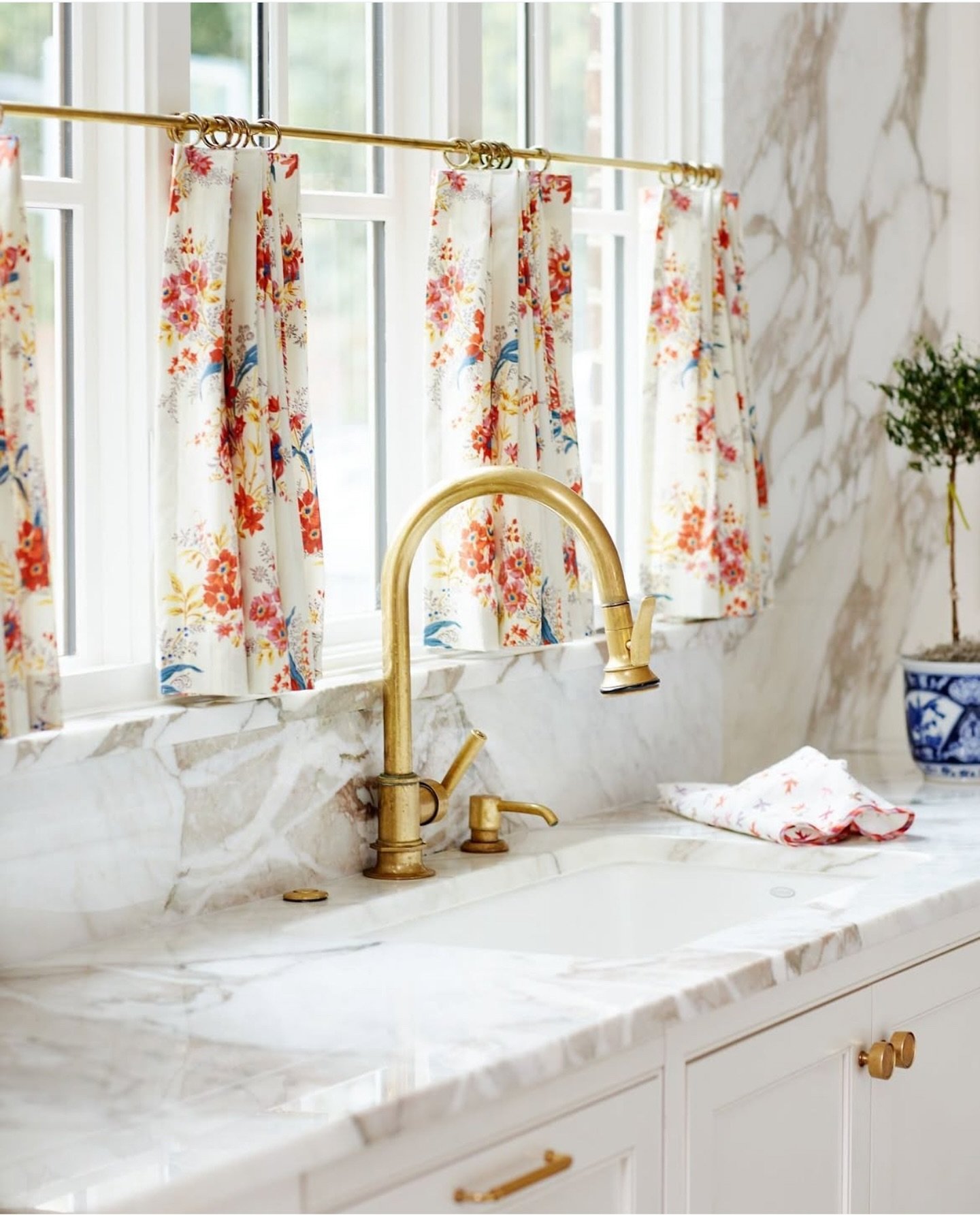White kitchen sink area with a brass gooseneck faucet, featuring short cafe curtains in a vibrant red and blue floral print hung on a brass ring rod over a marble counter.