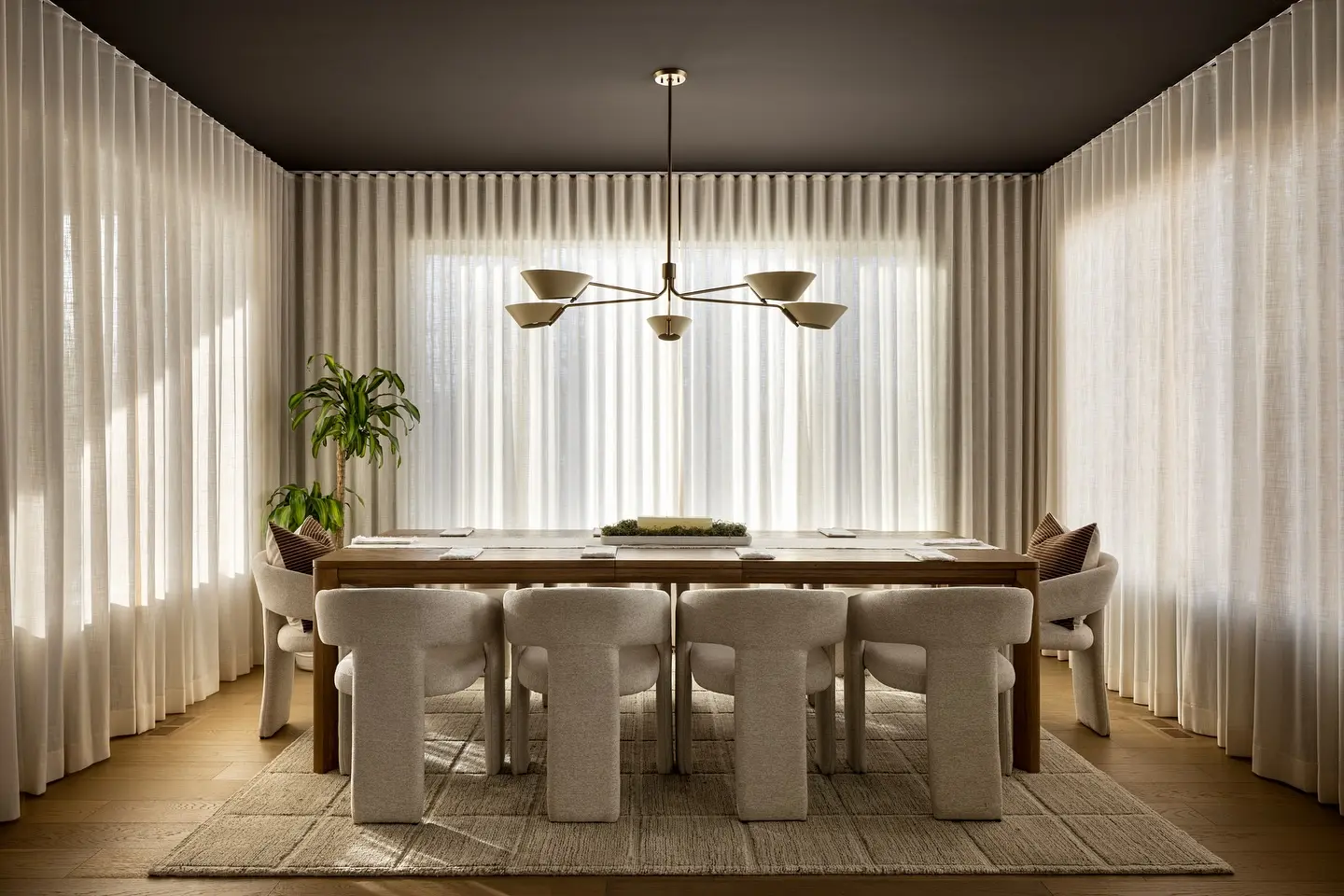 A minimalist dining room with a dark ceiling, soft sheer drapery surrounding the walls, a long wooden table, and plush, curved cream dining chairs.