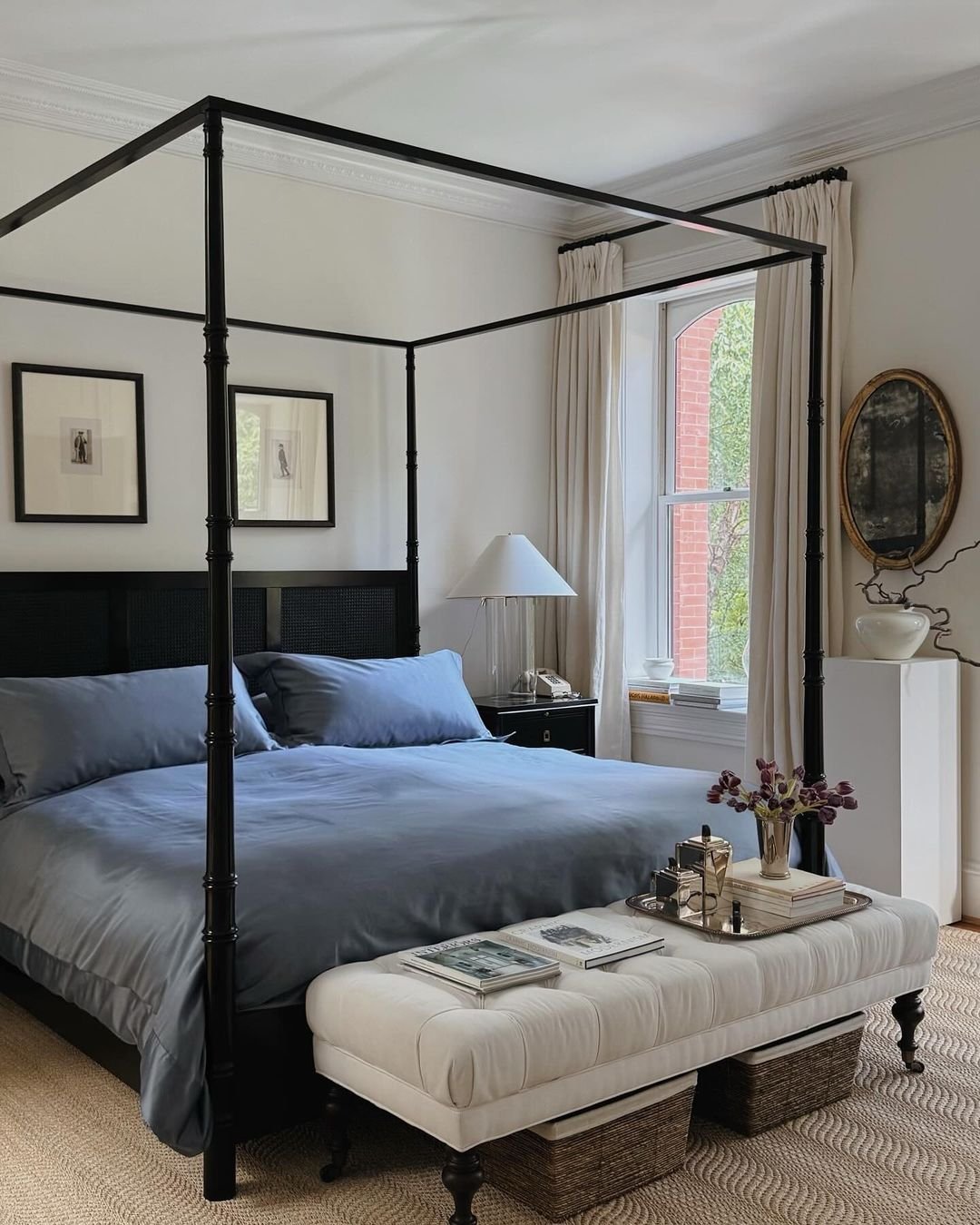 A formal bedroom with a black canopy bed, dusty blue bedding, and a long, white, tufted bench with turned legs at the foot of the bed.