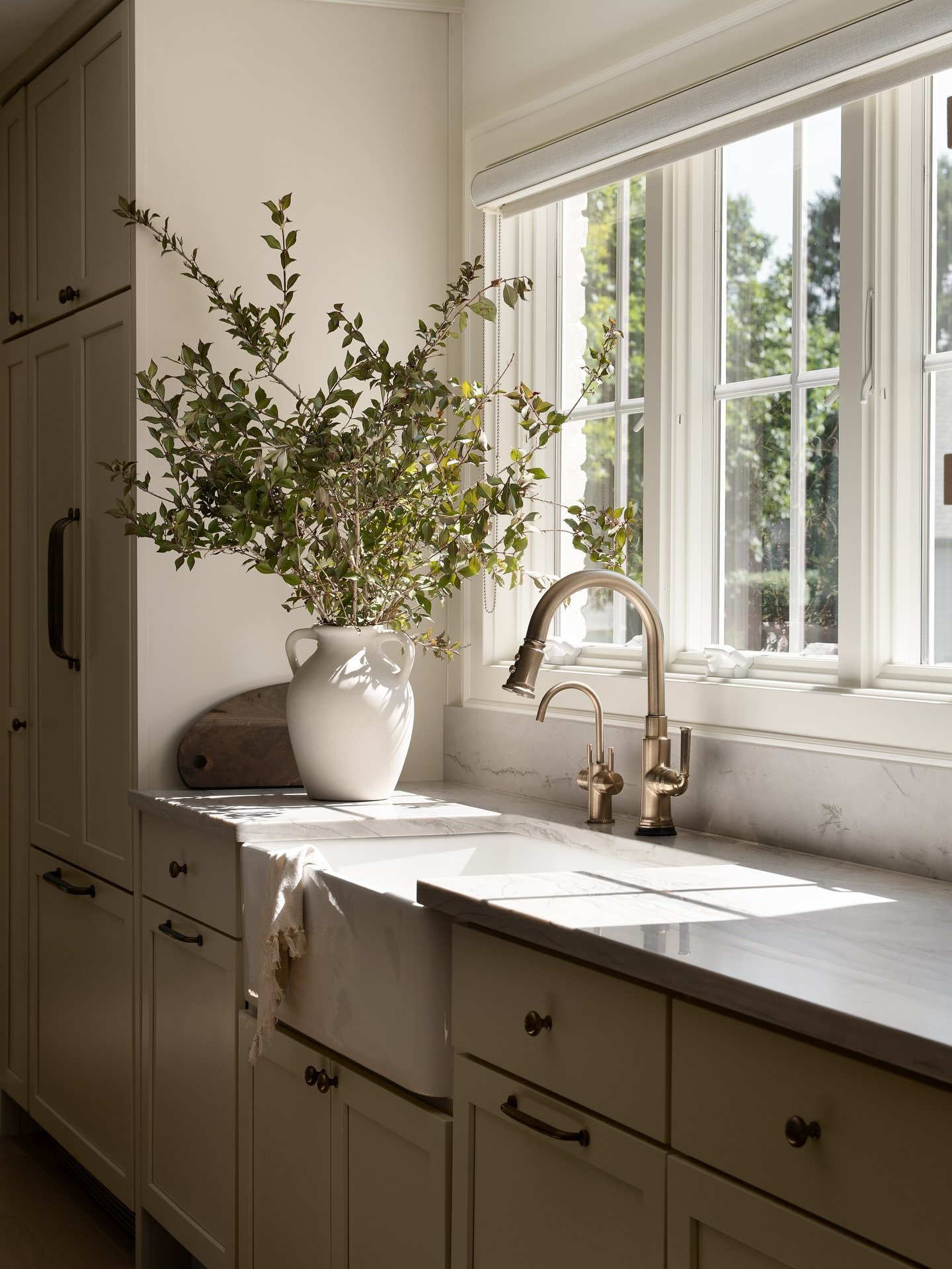 Kitchen sink area with creamy white cabinets, a farmhouse sink, and a brass faucet set against a marble countertop. A large grid window is covered by a pale, partially lowered Roman shade.