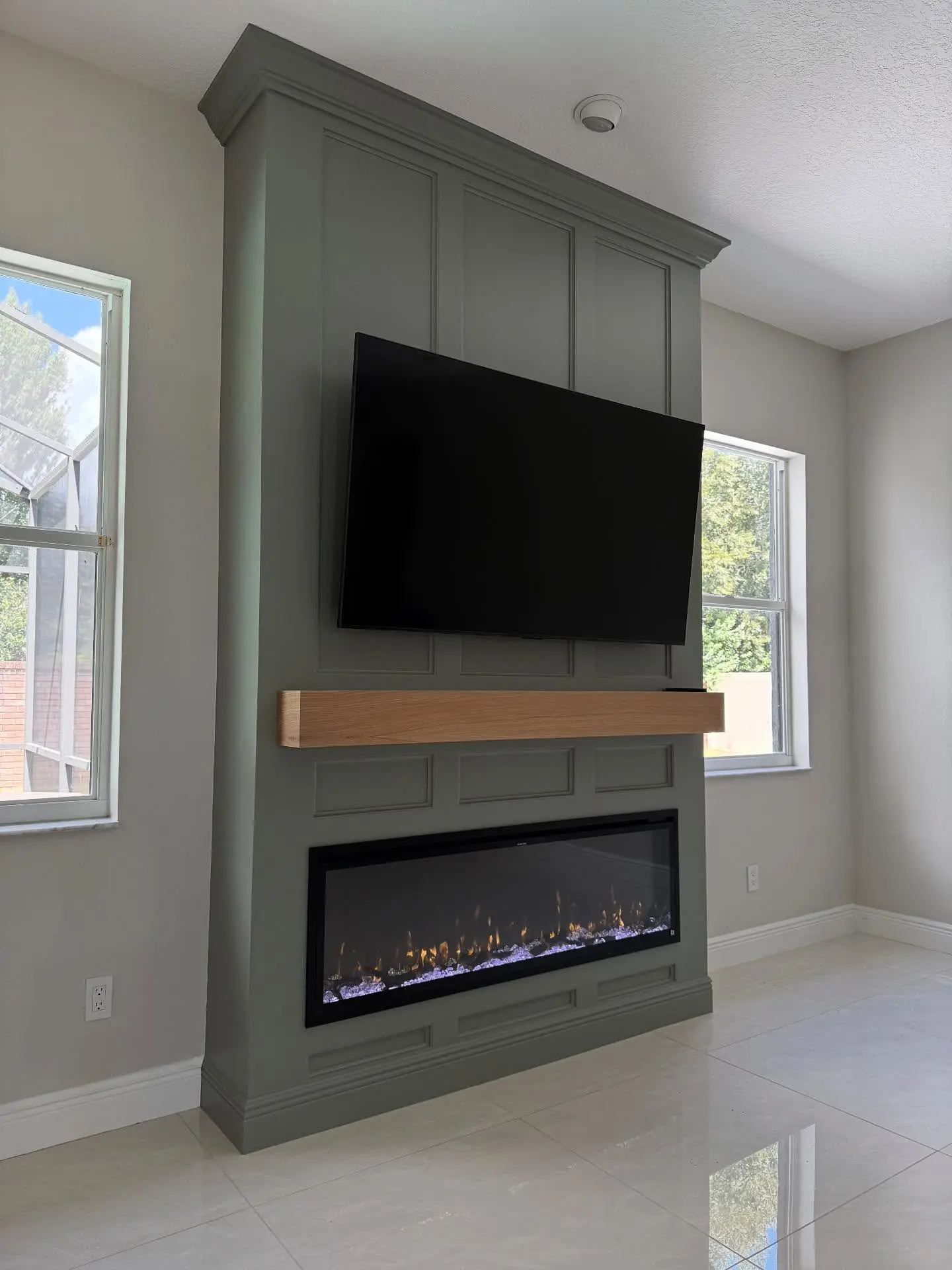 Modern floor-to-ceiling fireplace feature wall painted deep sage green with detailed paneling, a floating oak mantel, a wide electric fireplace, and a mounted TV.