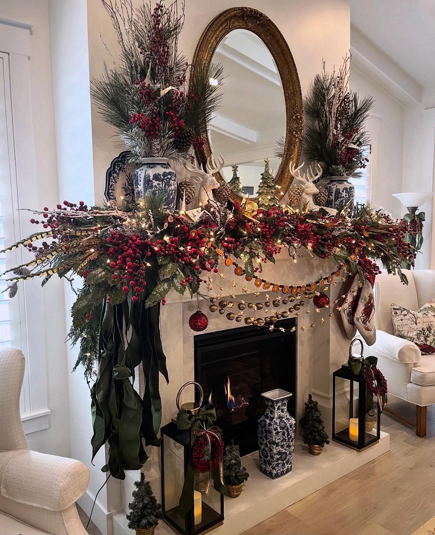 White stone fireplace mantel with an oval gold mirror, covered in a thick garland featuring red berries, pine branches, and white ceramic reindeer figures.