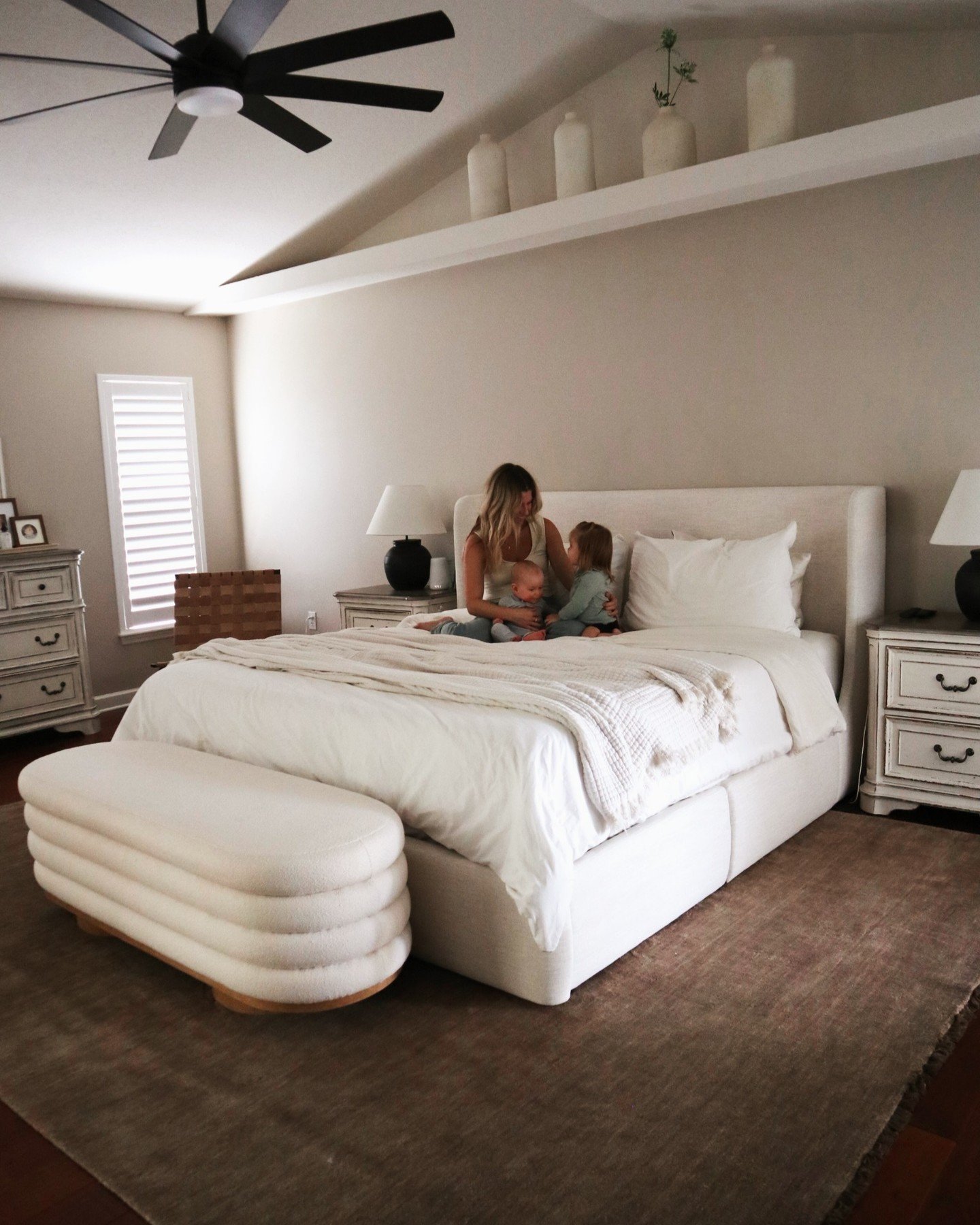 A bright bedroom with neutral walls, an upholstered white bed, and a thick, horizontally-layered white bouclé bench at the foot of the bed. A mother and two children are sitting on the bed.
