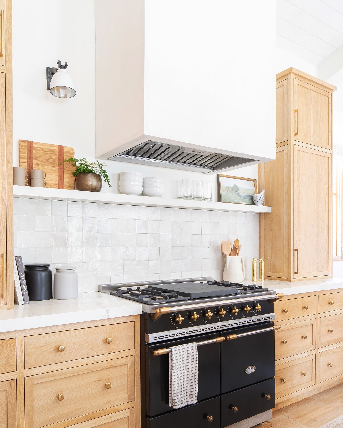 Contemporary kitchen with light wood cabinets, a black and brass range, a white square Zellige-style tile backsplash, and a floating shelf above the counter.
