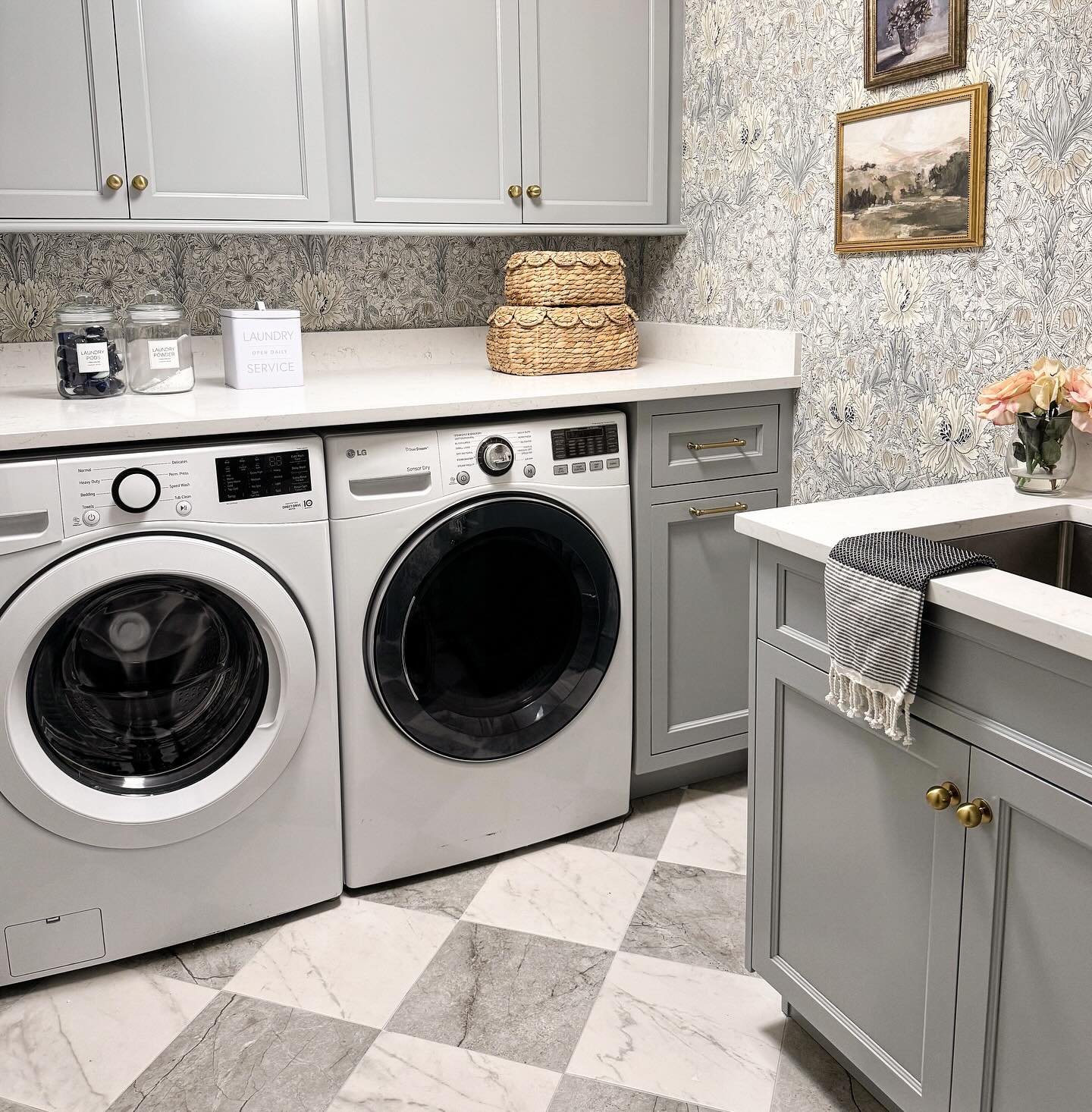 A small, charming laundry room with light gray cabinetry, a delicate floral wallpaper, and a striking gray and white checkerboard-patterned floor.