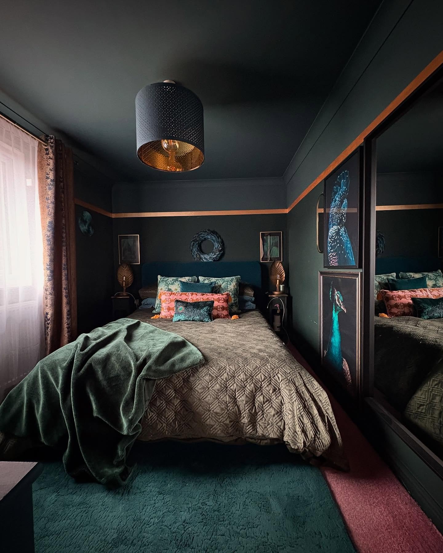Dark bedroom painted in deep forest green, featuring a quilted olive bedspread, peacock-themed art, mirrored wardrobes, and an eclectic mix of jewel-toned textiles.