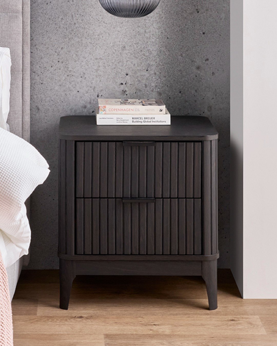 Dark charcoal or black nightstand with two drawers featuring a vertical fluted wood texture against a concrete-look wall.