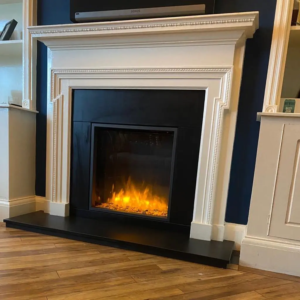 White traditional fireplace mantel and hearth against a dark navy blue wall, surrounding a recessed black electric fireplace insert with bright orange flames.