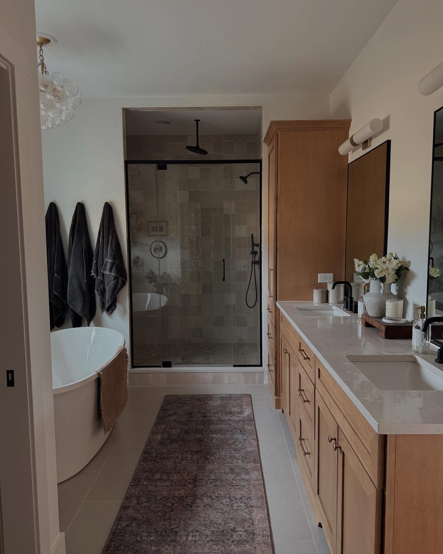 A contemporary master bathroom featuring light wood cabinetry, a freestanding tub, a walk-in shower with square tiles, and a long rectangular runner rug with a muted mauve and gray pattern.