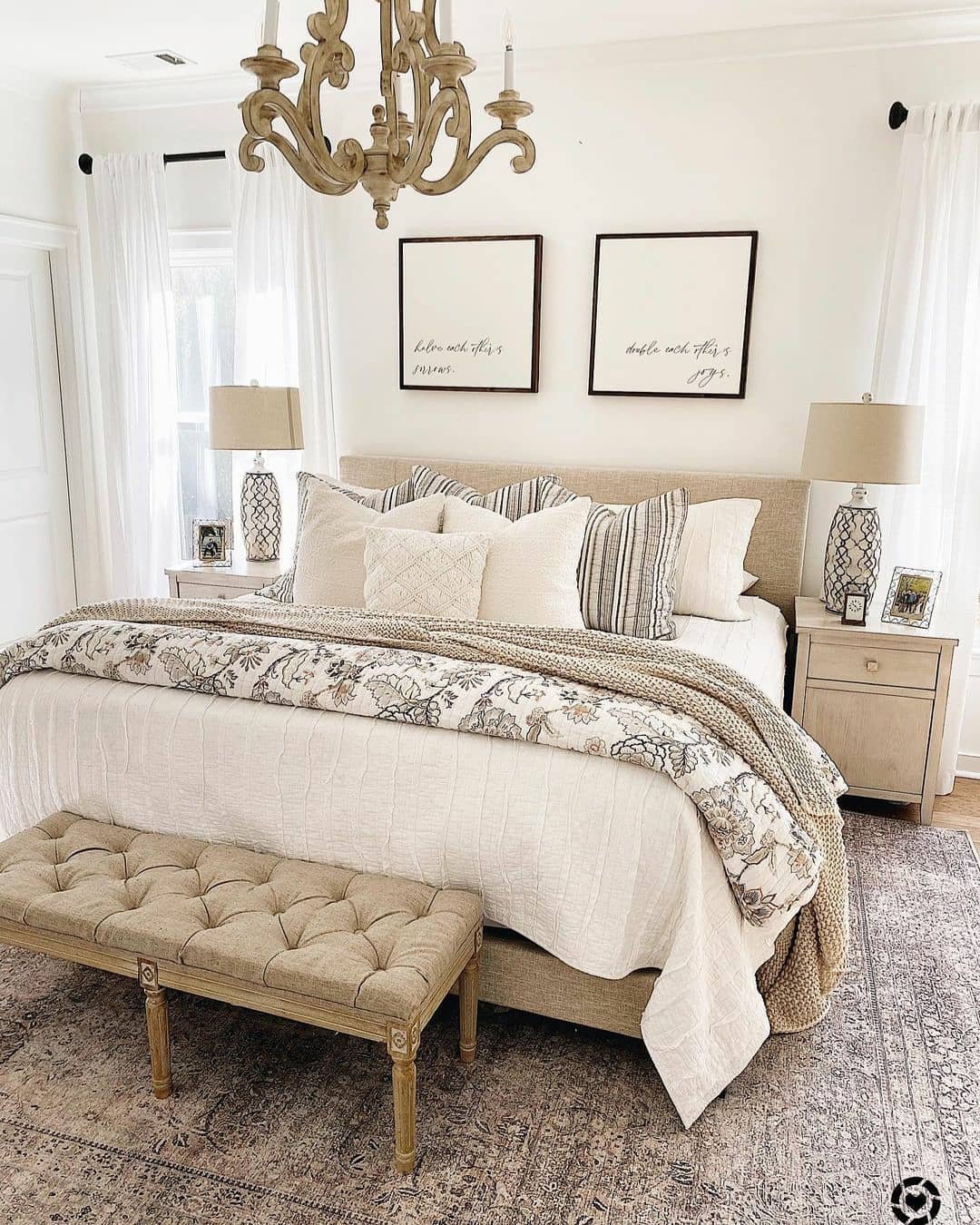 A luxurious beige master bedroom featuring a king-sized bed with layered bedding, a floral patterned quilt, a tufted upholstered bench, two large framed calligraphy prints, and a decorative wooden chandelier.