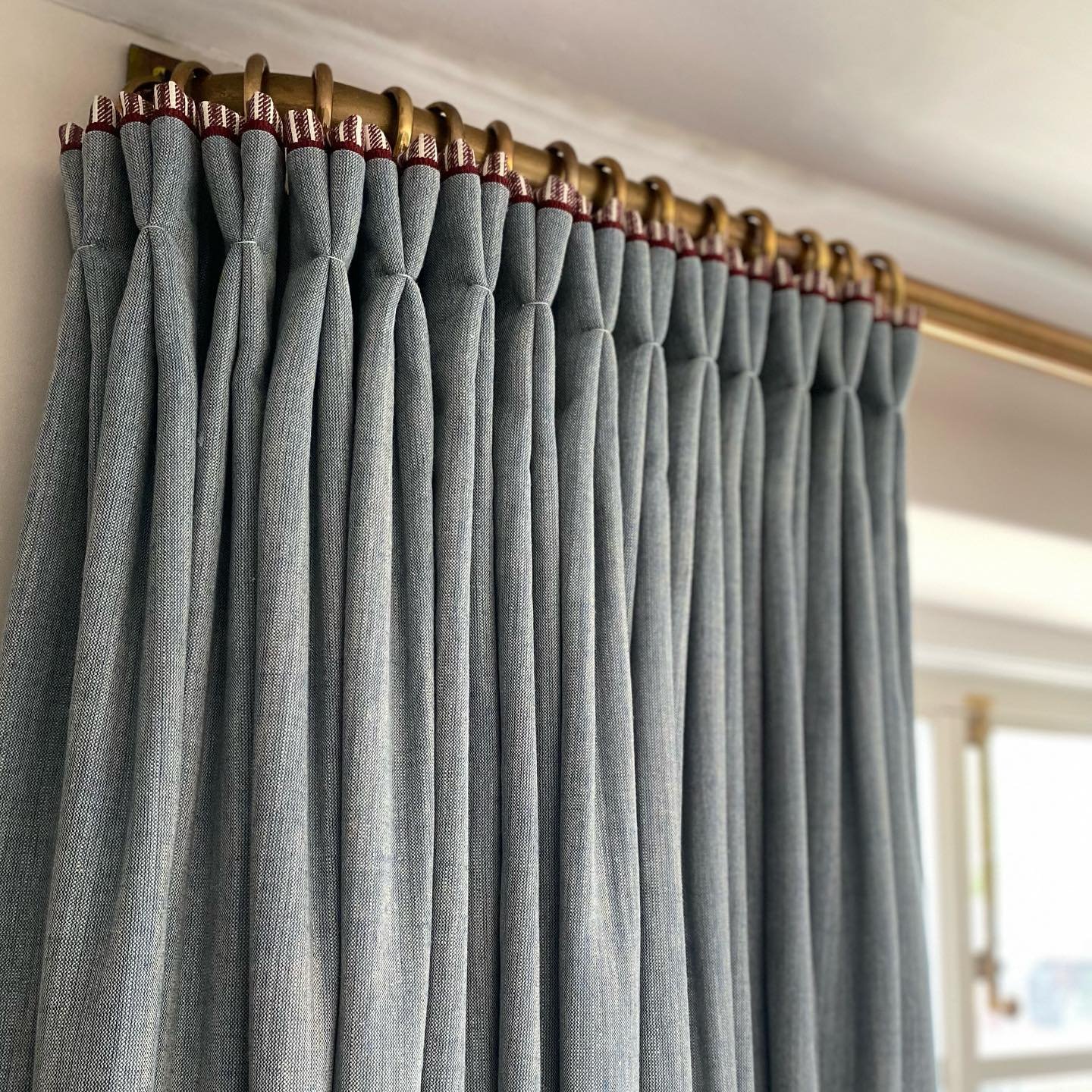 Close-up of tailored French pleated blue-gray curtains with a contrasting trim, hung on a polished brass curtain rod.