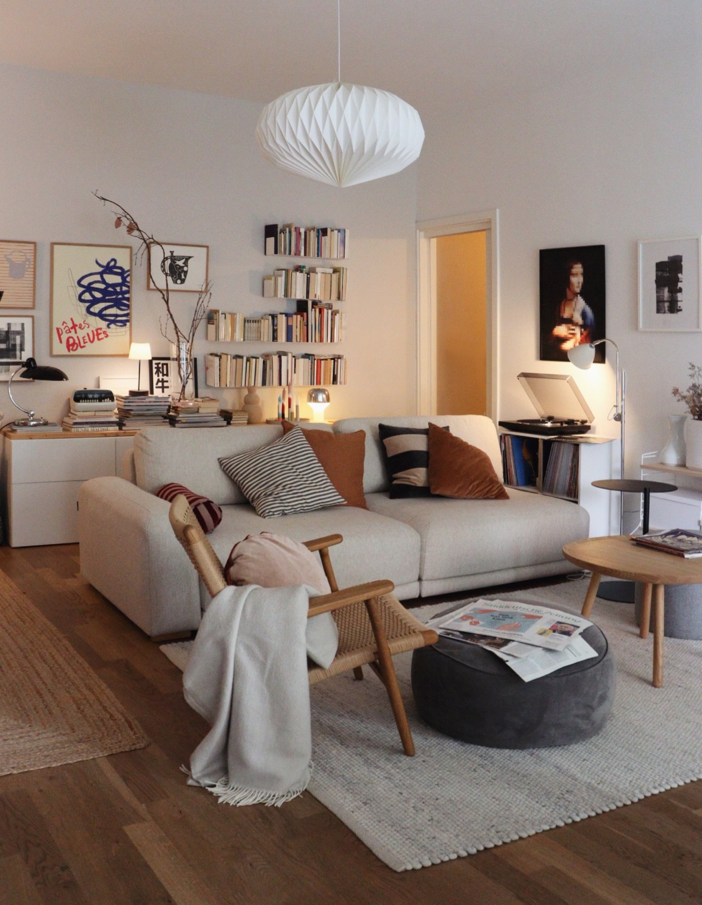 A small, bright living room featuring a compact light beige sofa, a low-hanging white paper lantern light, floating bookshelves, and a gallery wall above a side table.