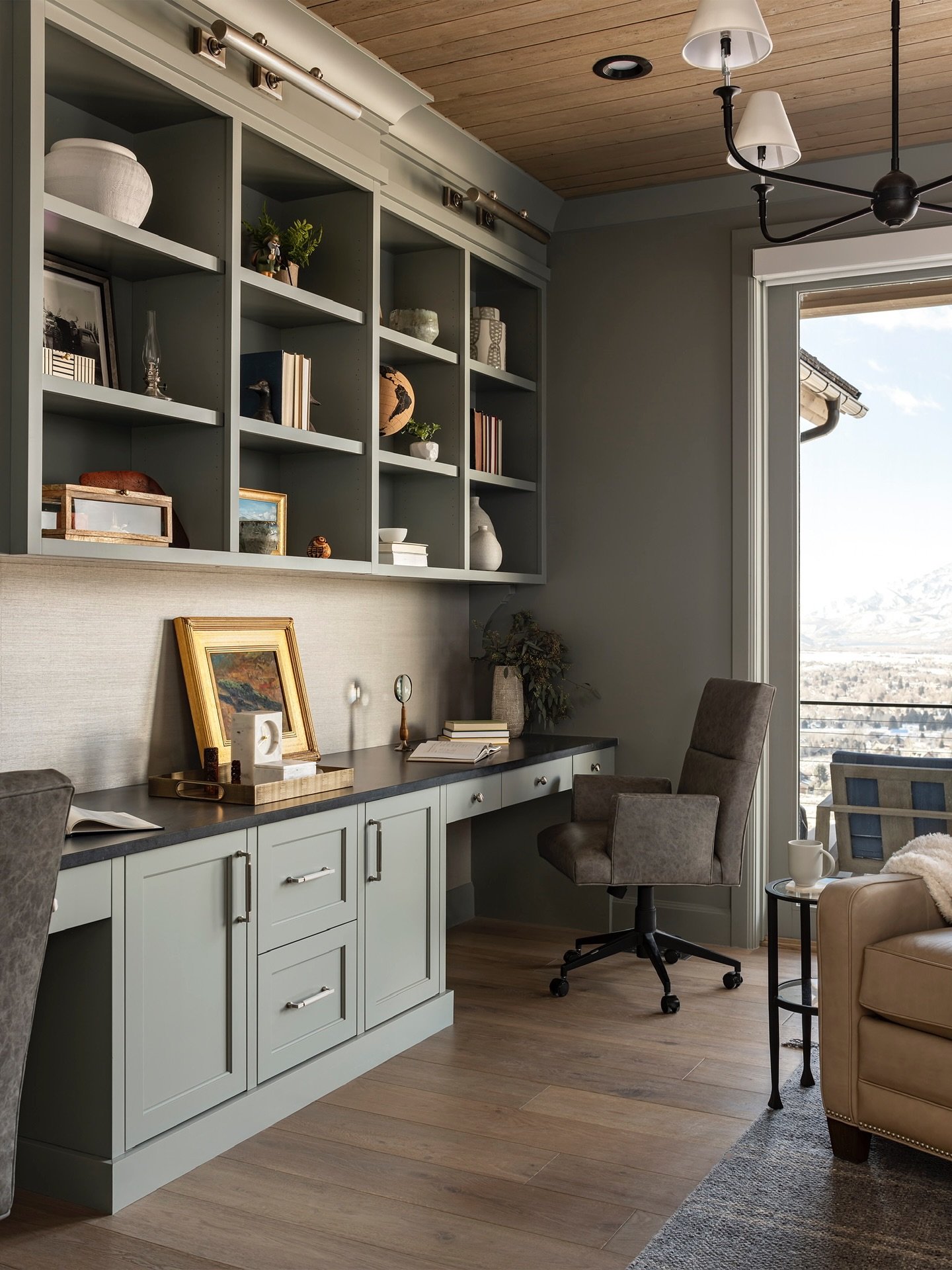 Sophisticated home office with dusty gray-green built-in desks and shelving, dark countertops, a gray rolling chair, and a view of a mountain landscape.