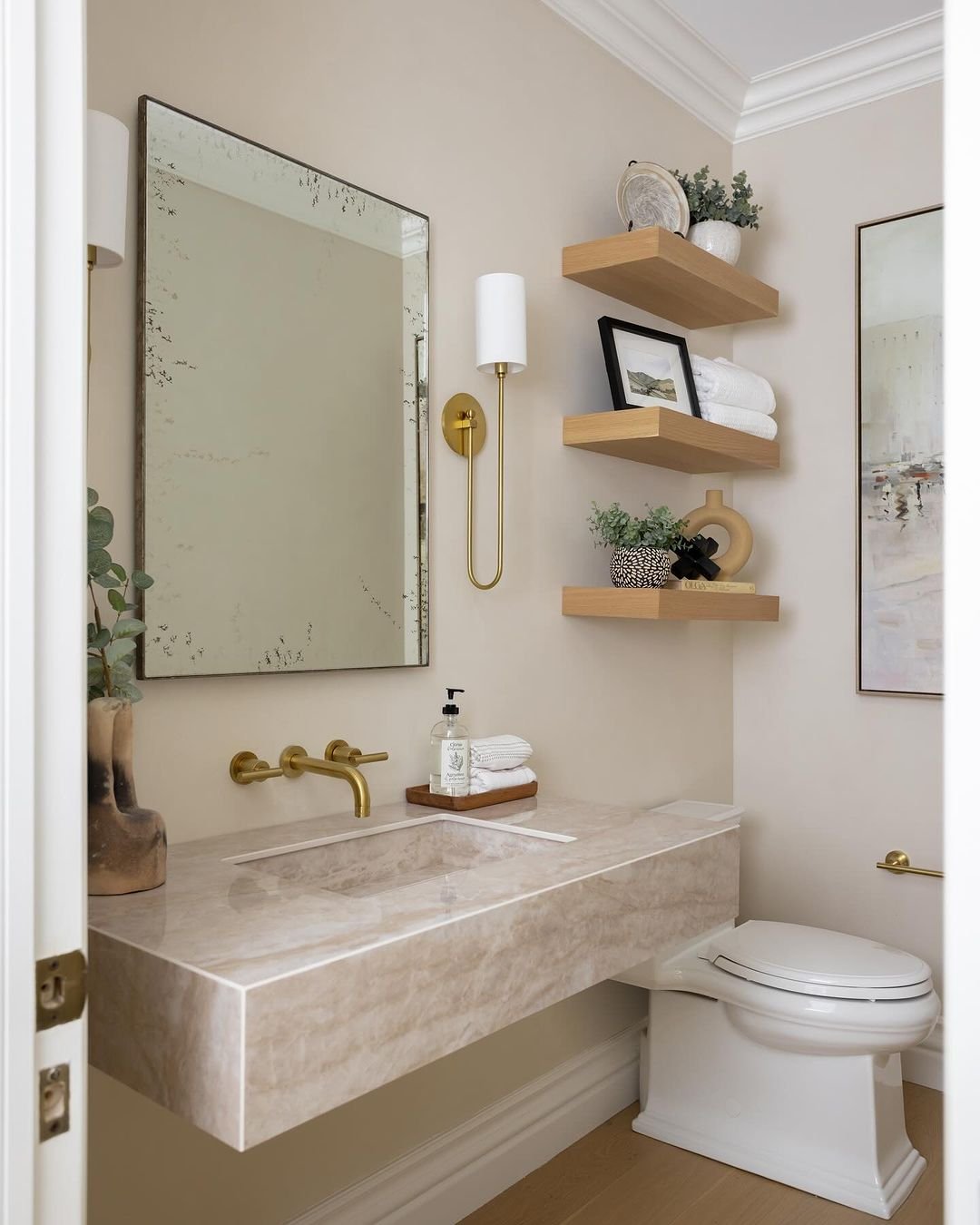  Three simple, thick light wood floating shelves stacked vertically next to a bathroom sink, styled with folded towels and small plants.
