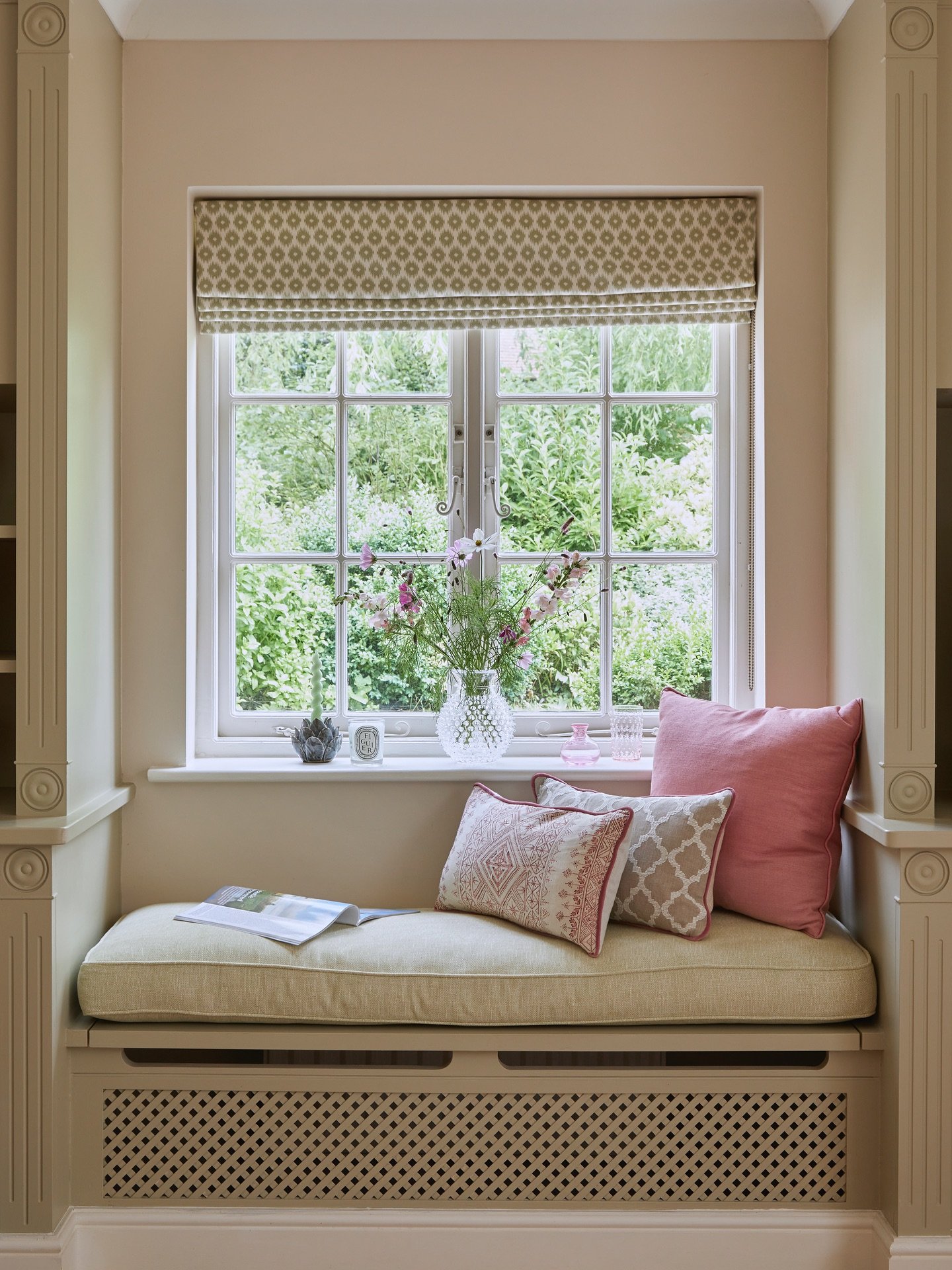 Elegant window seat built over a painted radiator cover, featuring a pale green cushion, pink and patterned throw pillows, and a geometric Roman shade.