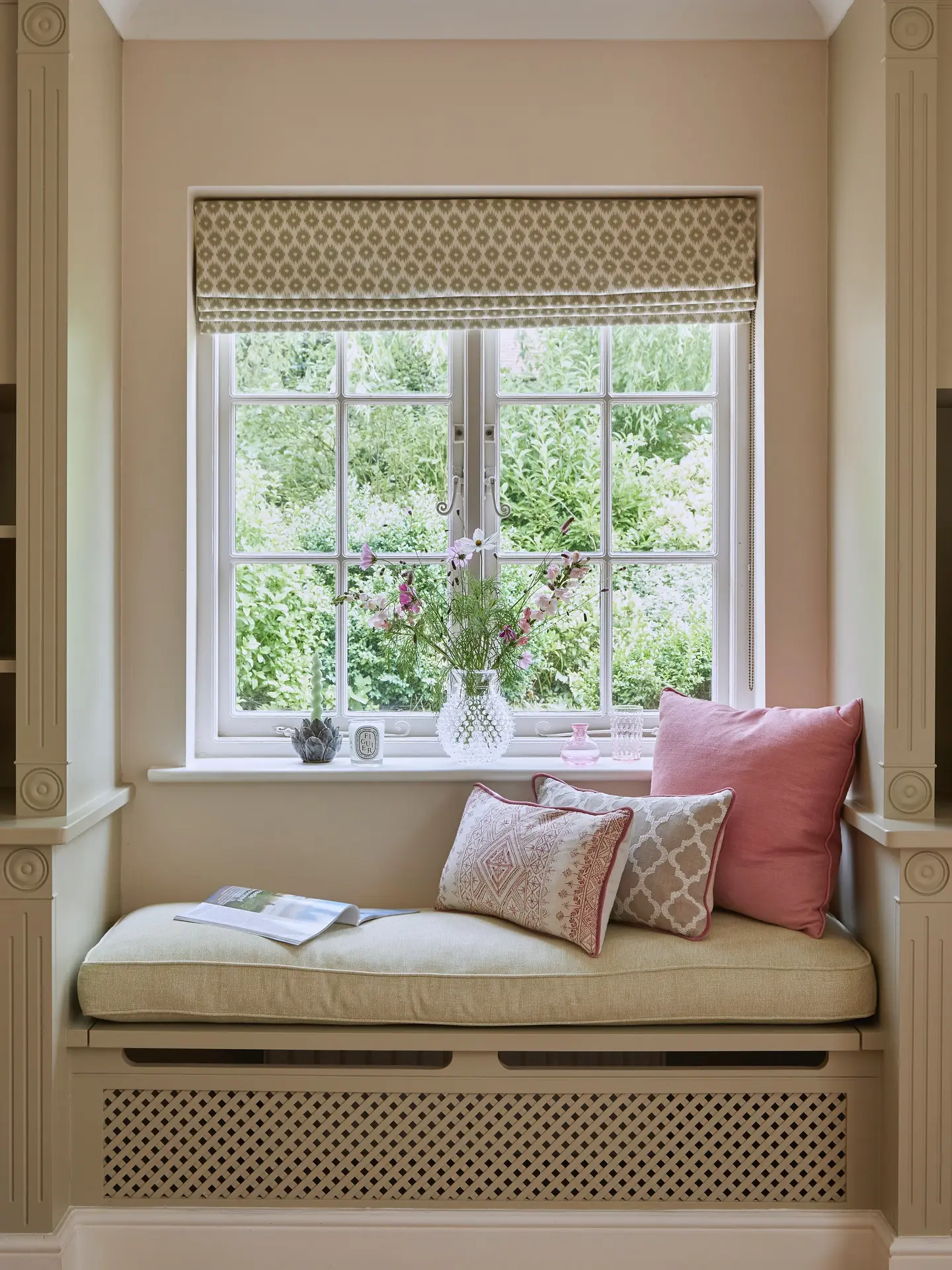 Elegant window seat built over a painted radiator cover, featuring a pale green cushion, pink and patterned throw pillows, and a geometric Roman shade.