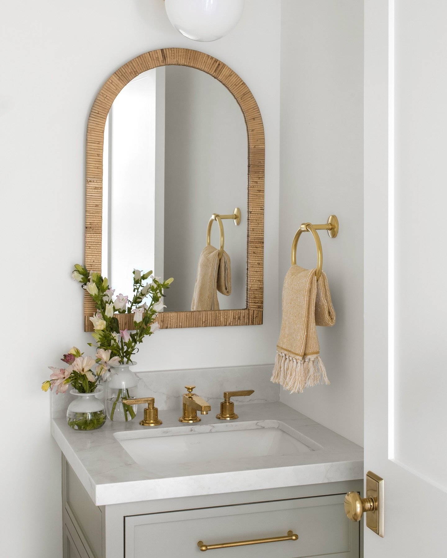 Light and bright powder room with white walls, a gray vanity, and a prominent arched mirror framed in natural rattan.