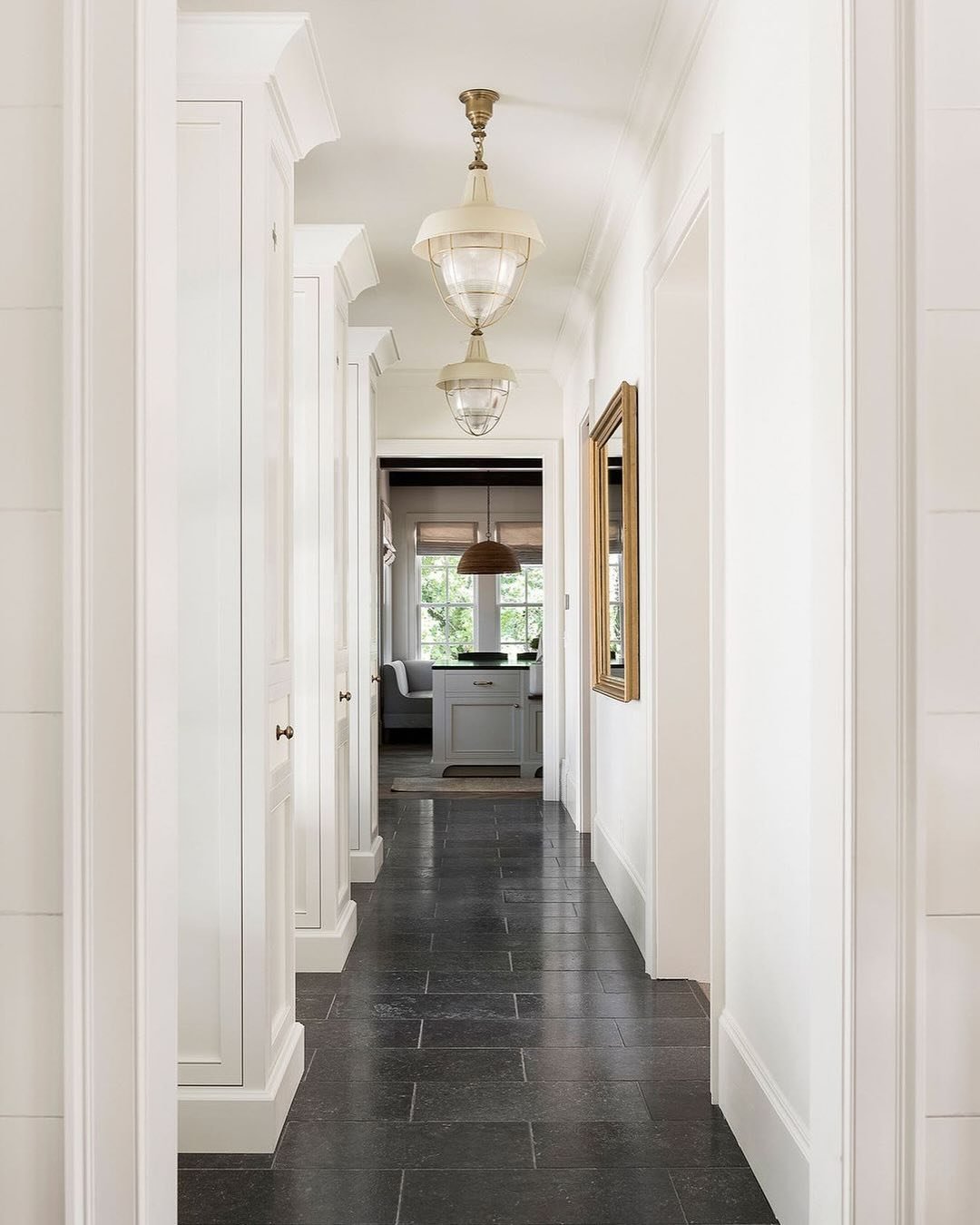White, narrow hallway with built-in cabinetry, dark slate floor tiles, and two ivory metal and clear glass industrial pendant lights.