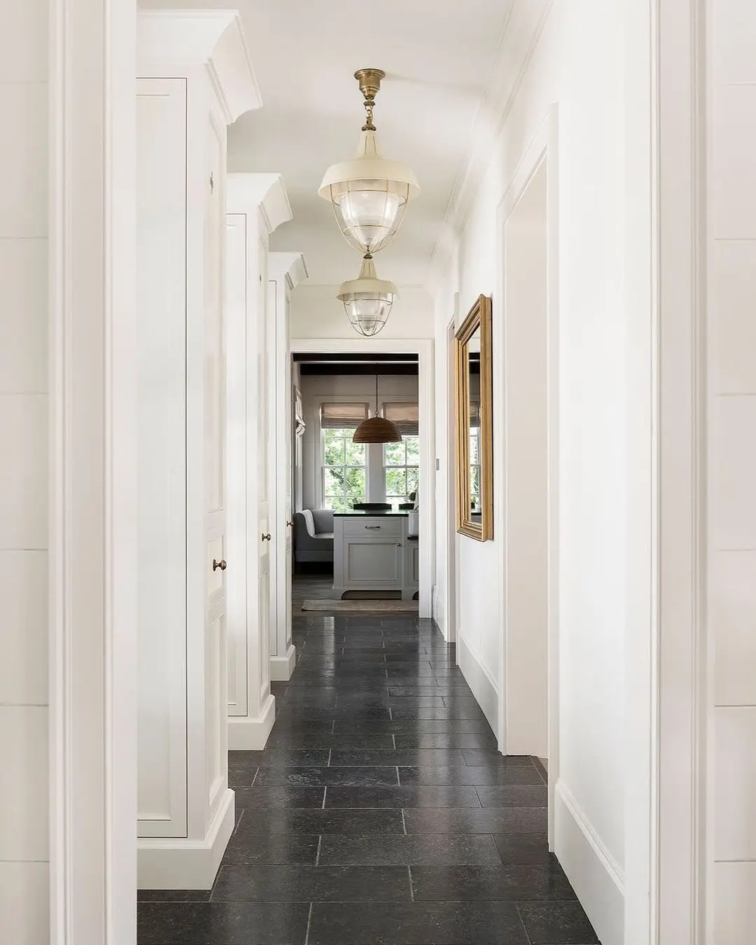 White, narrow hallway with built-in cabinetry, dark slate floor tiles, and two ivory metal and clear glass industrial pendant lights.