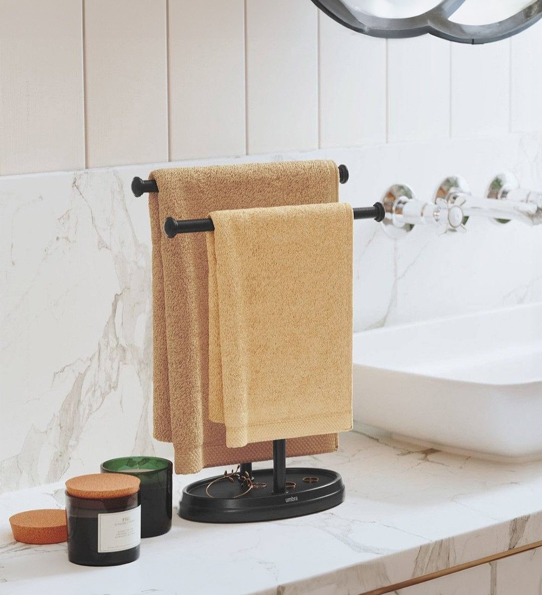A black metal free-standing countertop towel holder with two bars, holding two beige hand towels on a marble vanity.