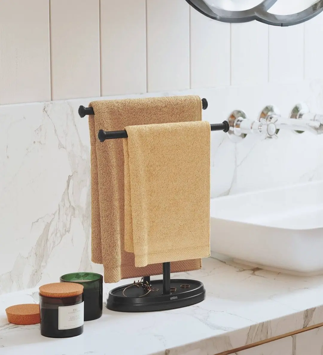 A black metal free-standing countertop towel holder with two bars, holding two beige hand towels on a marble vanity.