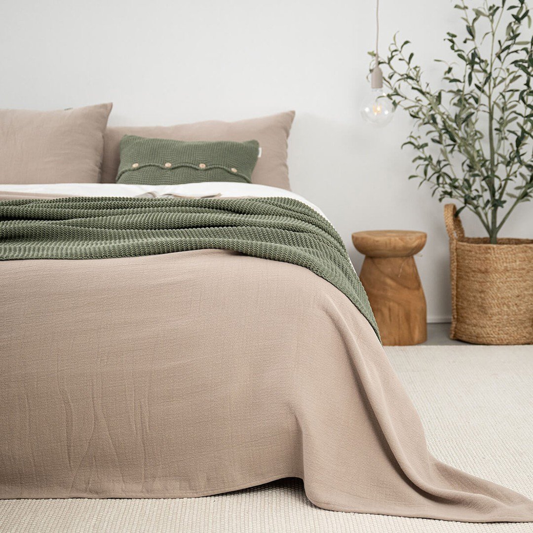 A close-up of a bed featuring a taupe bedspread, a chunky knitted olive green throw, and a small green bolster pillow.