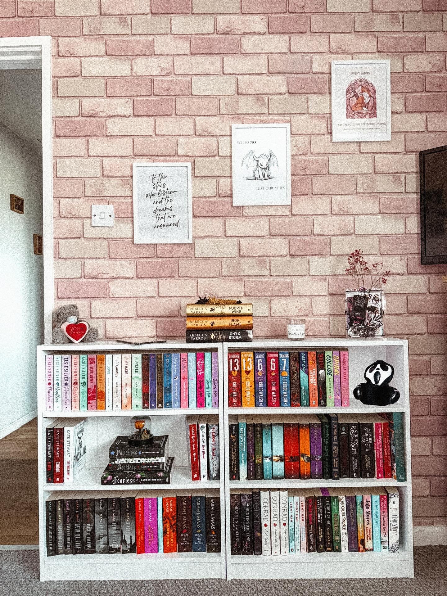 Two short, wide white cube shelving units placed side-by-side against a pink brick-patterned wallpaper, displaying books organized by color and small decorative items.