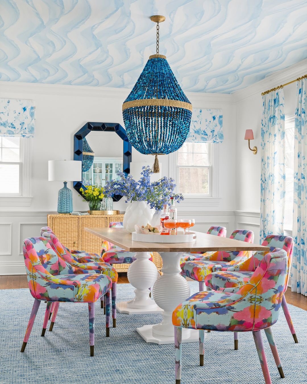 A bright, eclectic dining room with a blue watercolor ceiling and bold floral upholstered chairs around a light wood pedestal table, beneath a large blue beaded chandelier.