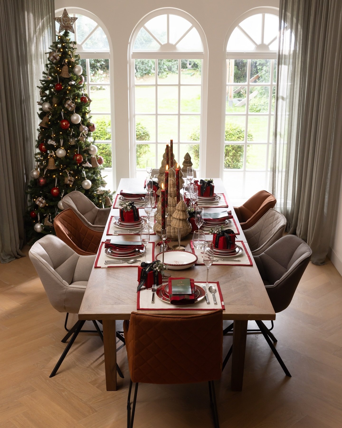 A stylish Christmas dining table with quilted dining chairs and a wood surface, featuring red placemats, layered dark and white plates, and a centerpiece of wooden conical Christmas trees.