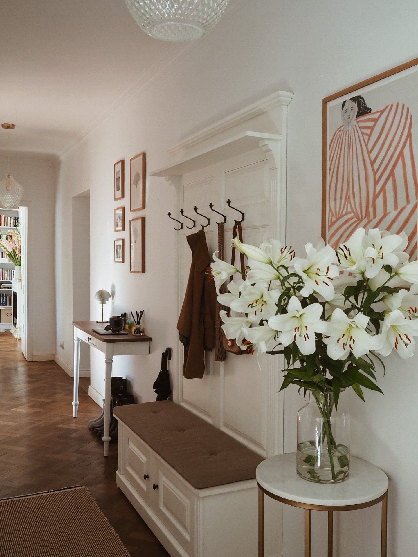 A white hallway with herringbone wood floors, a large white bench with coat hooks and a cushion, and a modern art print.